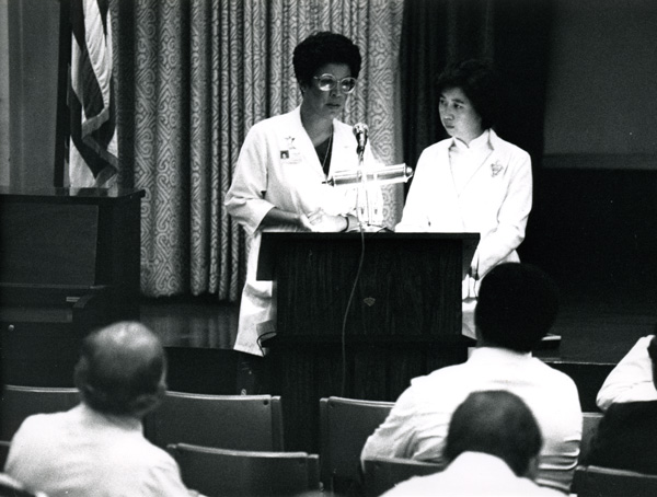 A woman in white medical coat standing at a lectern.