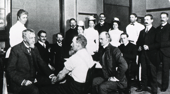 Dr. Mitchell examining a male patient in left foreground while a group of men and women look on.