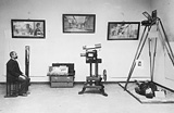 Historical photograph of a man sitting in a room with multiple mirrors and a complex camera equipment set-up