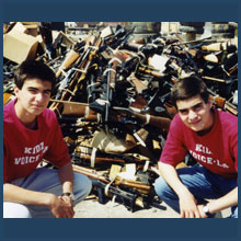 Niko and Theo Milonopoulos pose in front of large pile of guns