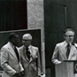 A black man talking with two white men next to a chalkboard and podium.