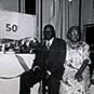 African American male and female sharing a seat in a room with presents and a long table. “50th” on the board behind the table.