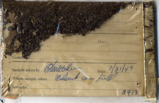 Clear plastic envelope containing a sample of dirt and a paper card with blue ink handwriting.