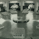 (upper)Trays of small bottles are on a large circular surface; workers are visible around the perimeter; (lower) worker scoops material into a small funnel