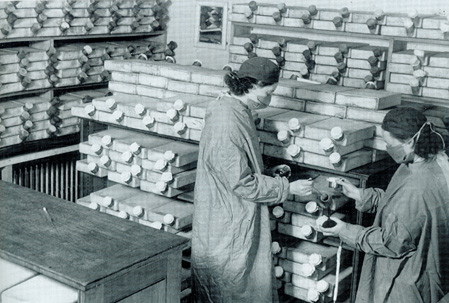 Two women in surgical gowns, masks, and head coverings in a room stacked with square ceramic penicillin vessels. 