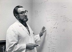 A black and white photograph of a man in a lab coat standing at a white board.