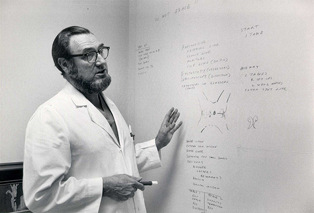 A black and white photograph of a man in a lab coat standing at a white board.