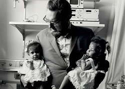 A black and white photograph of Koop in a suit holding the separated twins in street clothes.