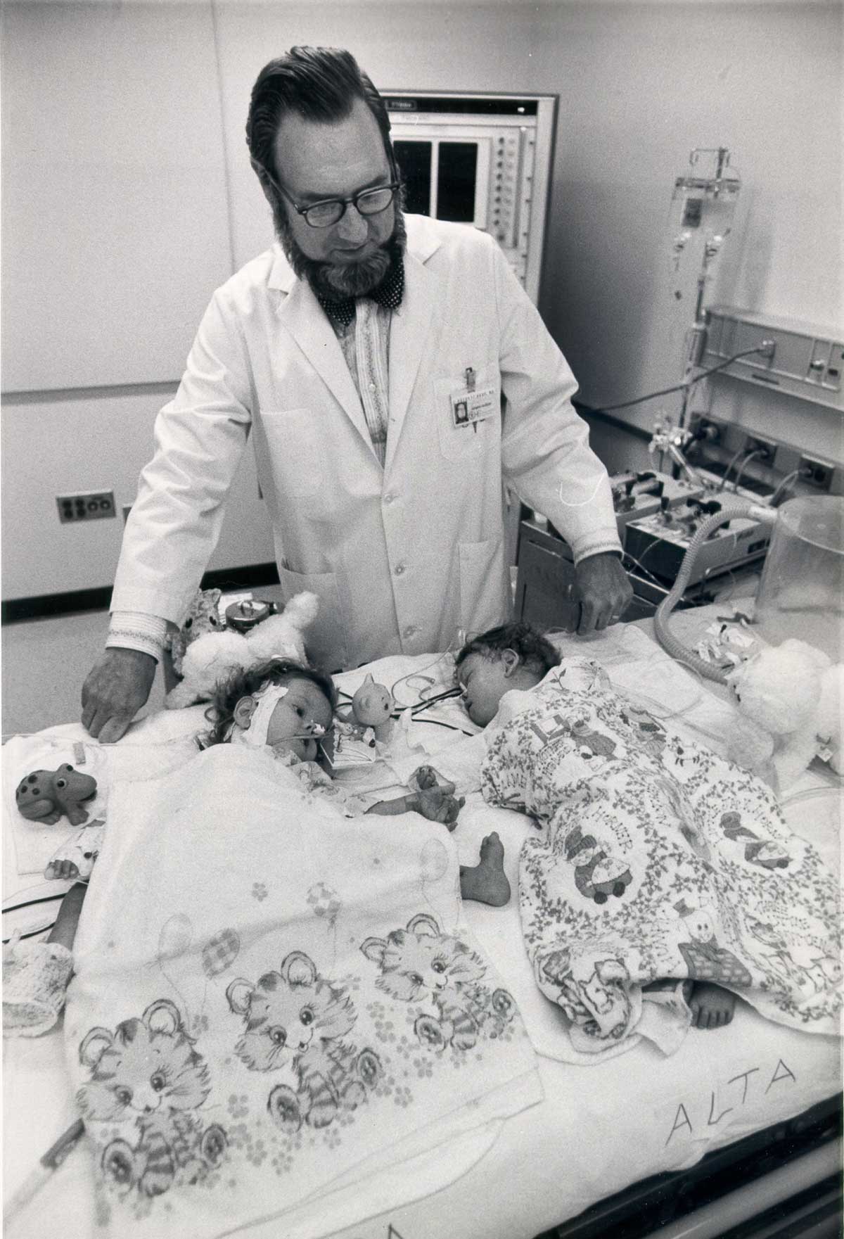 A black and white photograph of Koop in a lab coat by the twins lying under blankets.