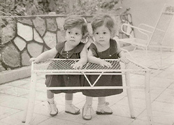 A photograph of the young twins standing together leaning on a coffee table outdoors.