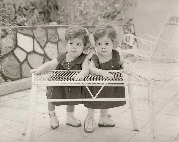 A photograph of the young twins standing together leaning on a coffee table outdoors.