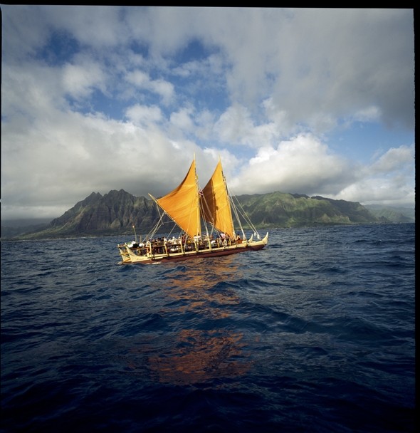 Color image of a double-hulled canoe with two orange sails at sea. An island can be seen in the distance.
